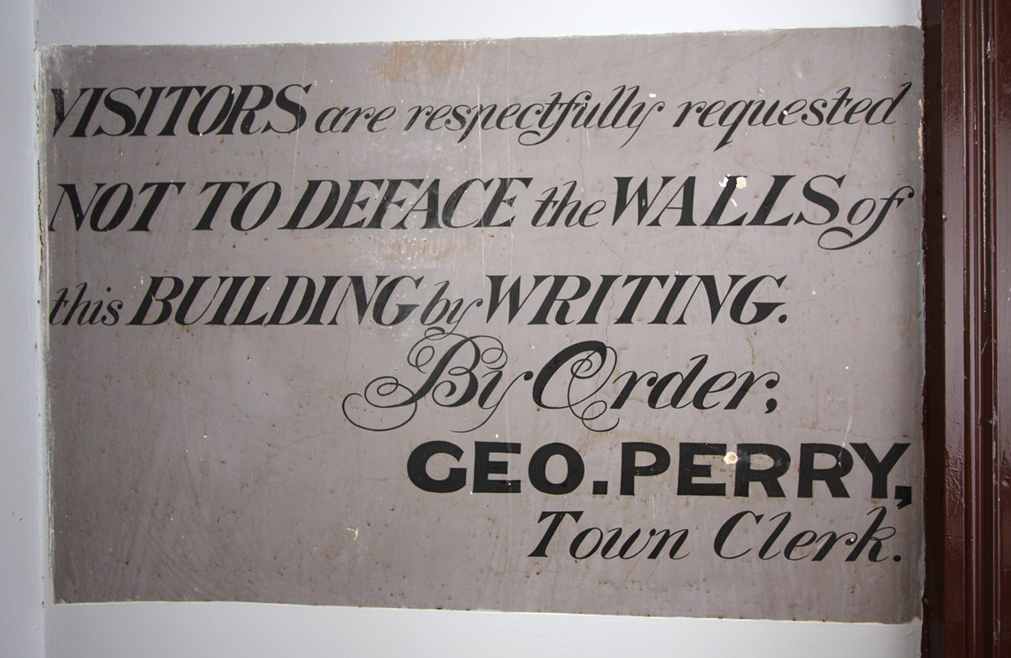 Painted sign that reads Visitors are respectfully requested not to deface the walls of this building by writing. By Order: CEO Perry, Town Hall. 